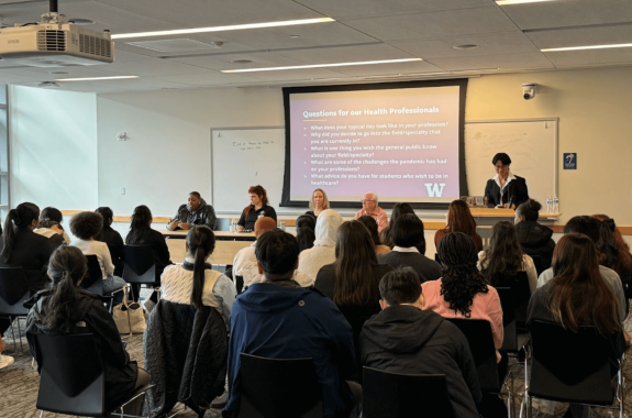 Panel Discussion Event: A group of students attentively listens to a panel of healthcare professionals seated at the front of the room. A presentation slide behind the panelists lists discussion questions, and one speaker stands at a podium.