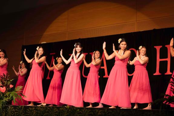 A group of women in pink dresses and adorned in lei perform hula on a stage in the HUB ballroom.