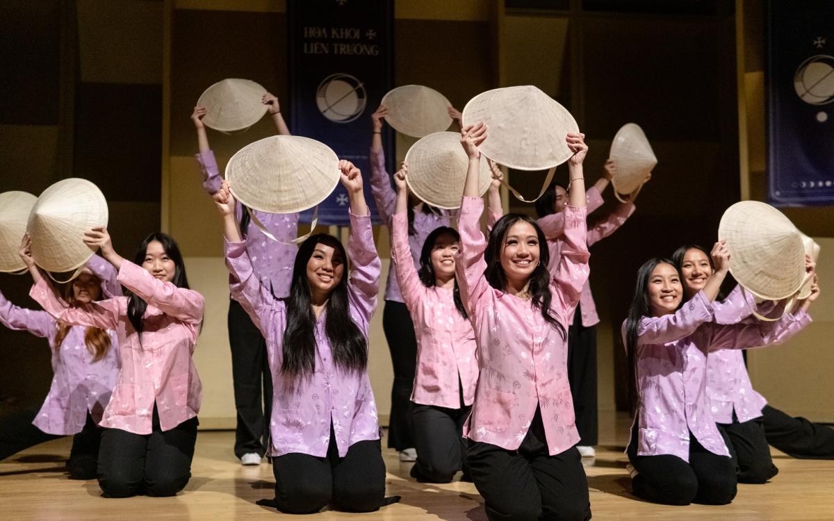 The pageant contestants dancing in Vietnamese traditional clothing while holding rice hats, smiling through their performance.  