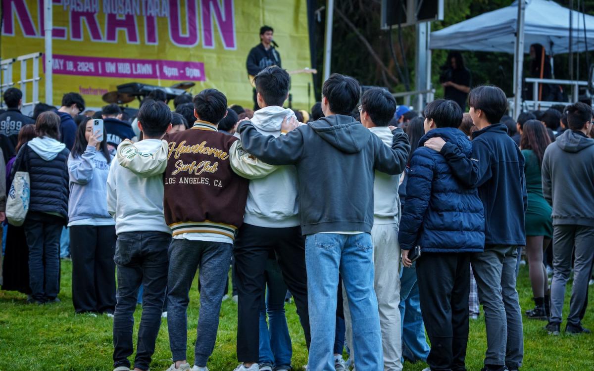 Group of students with arms on one another backs watching musician on stage performing