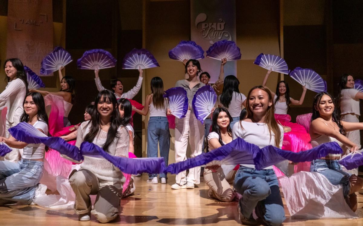 Shows 20 dancers on stage smiling during the ending pose, holding traditional purple short fans and pink long fans. 