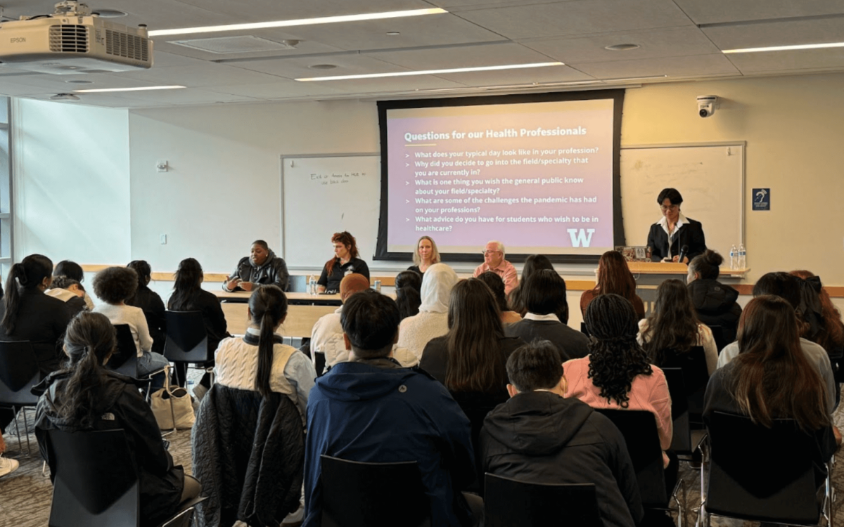 Panel Discussion Event: A group of students attentively listens to a panel of healthcare professionals seated at the front of the room. A presentation slide behind the panelists lists discussion questions, and one speaker stands at a podium.