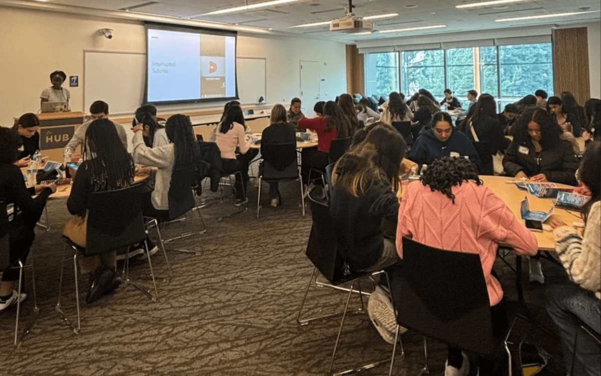 Interactive Workshop Setting: A classroom-style workshop with several tables, where students are engaged in hands-on activities. A speaker stands at the podium next to a large screen with the title "Interrupted Sutures".