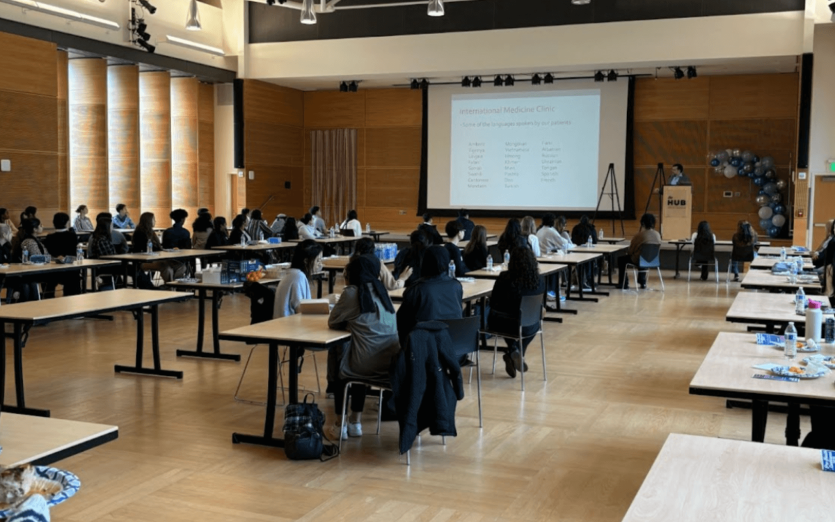 Large Presentation Session: A spacious event hall where students are seated at long tables, facing a large presentation screen. The slide displays information related to an "International Medicine Clinic," discussing multilingual patient care.