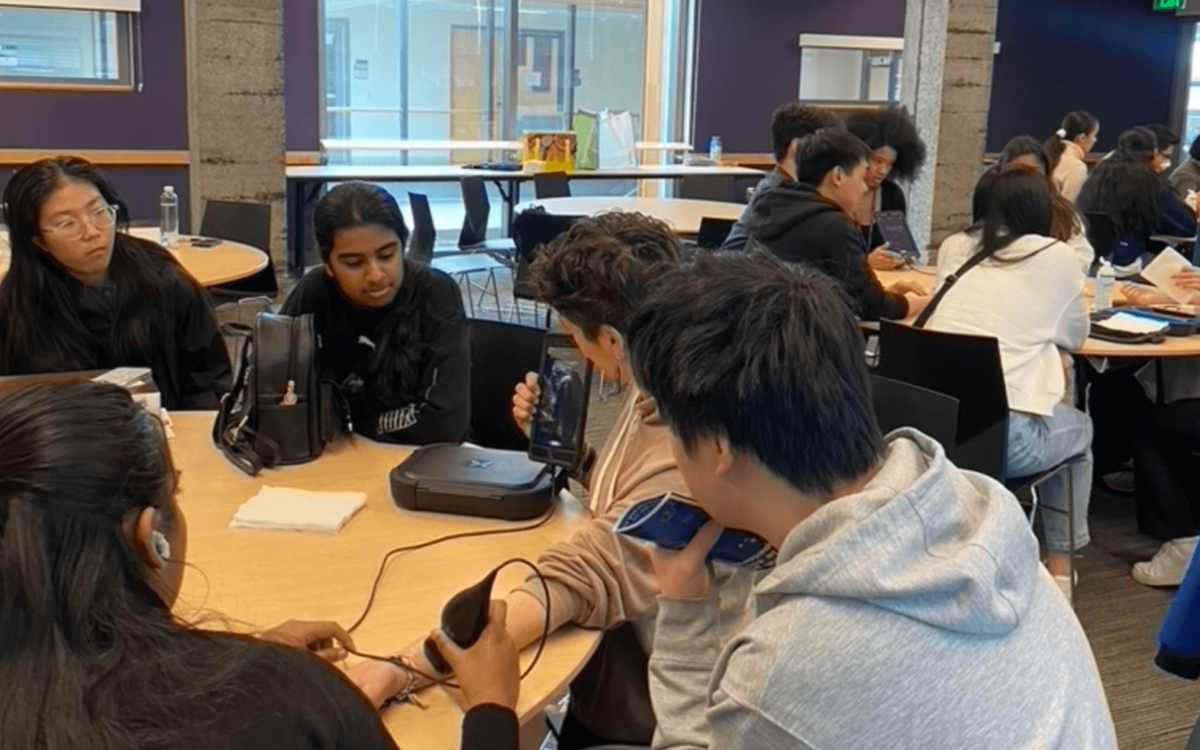 Hands-On Medical Training: A group of students gathered around a table, using medical devices to perform a task. One person holds a Doppler ultrasound wand on a peer's arm, while others observe a connected tablet displaying the ultrasound image.