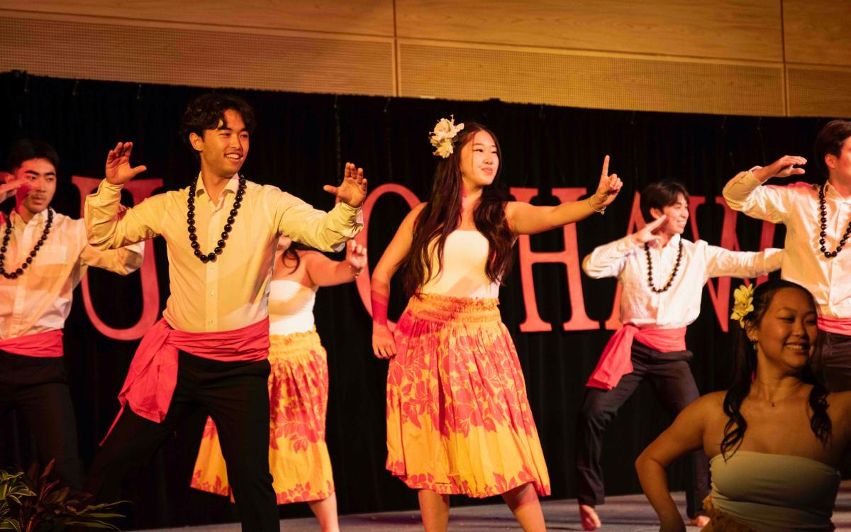 Pairs of students in traditional outfits dance hula on the HUB ballroom stage.