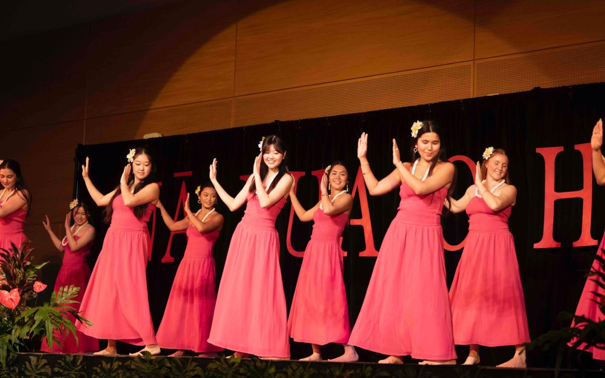 A group of women in pink dresses and adorned in lei perform hula on a stage in the HUB ballroom.